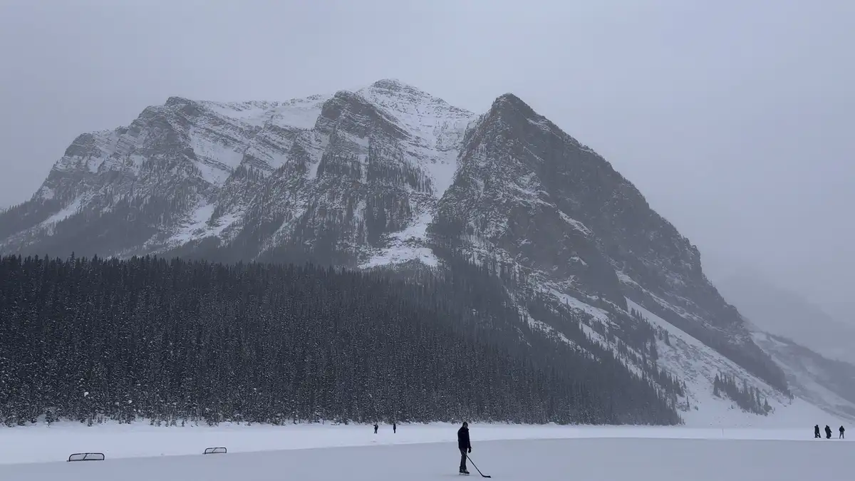 A person stands alone on a vast frozen lake, holding a hockey stick, with snow-covered mountains and a dense forest in the…