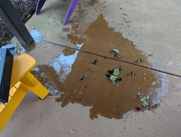 A close-up photo shows a brown liquid spill on a concrete patio, with shattered glass fragments scattered across the wet…
