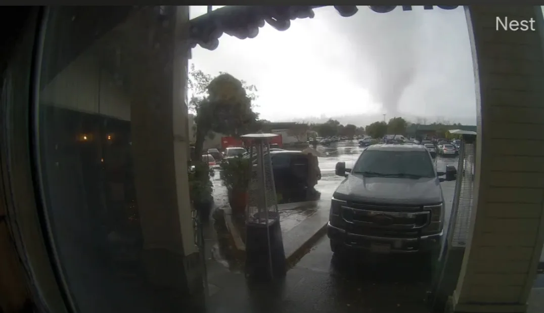 A fisheye security camera view from inside a building looking out onto a wet parking lot where a large tornado is visible in…