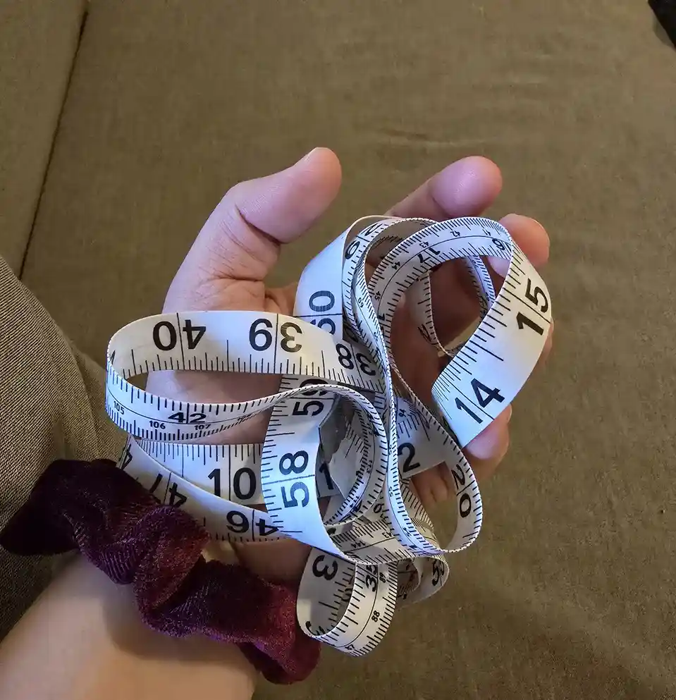 A person's hand holds a tangled white measuring tape with black numbers and markings, showing various sizes like 14, 15, 38…