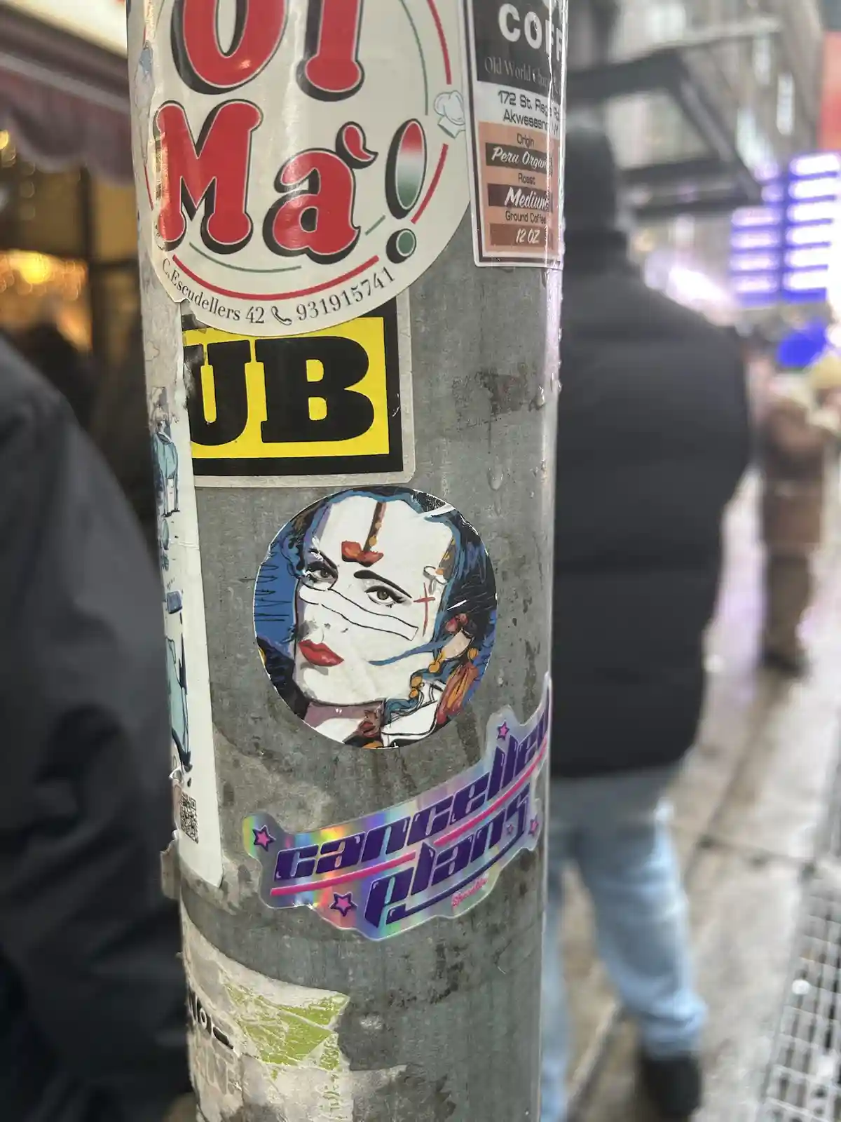 A close-up of a metal pole covered in various stickers, including a prominent holographic one at the bottom that reads…