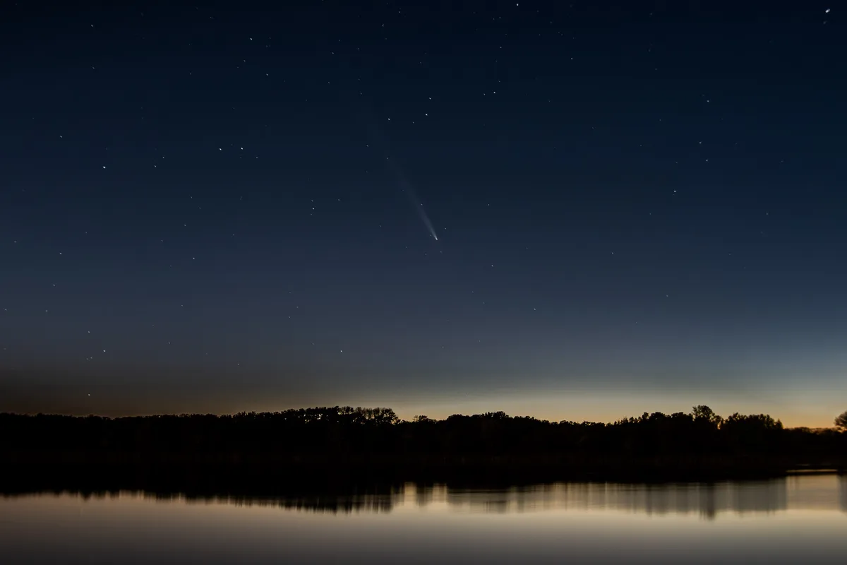 A serene night sky over a calm body of water reflects the dark silhouettes of trees along the horizon. A bright comet with a…