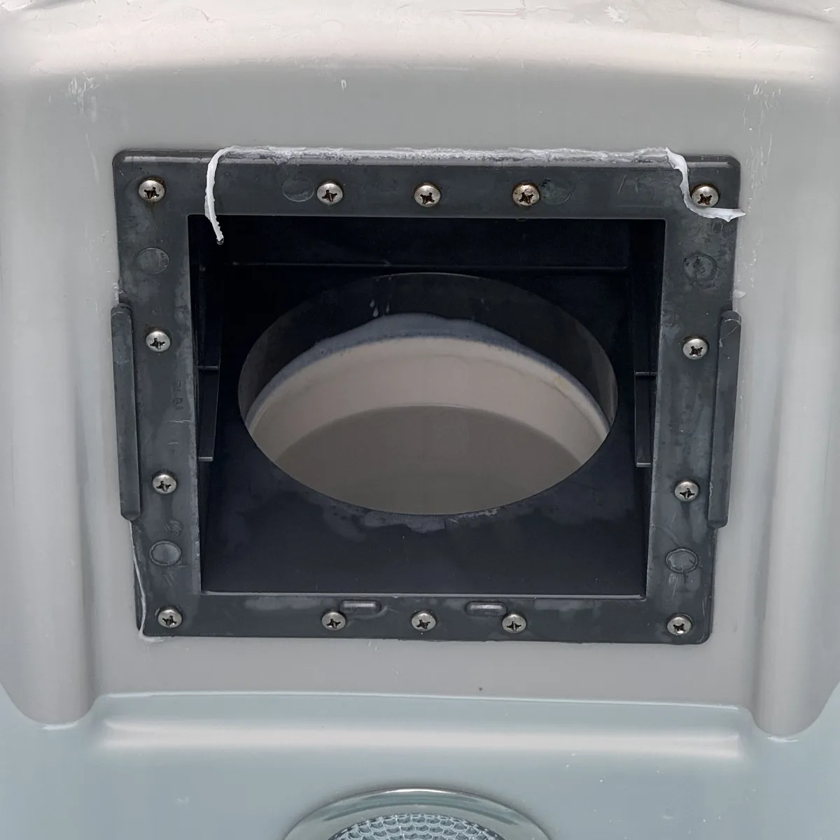 A close-up photo shows a square metal frame screwed into a light gray plastic surface, revealing a circular white ceramic…