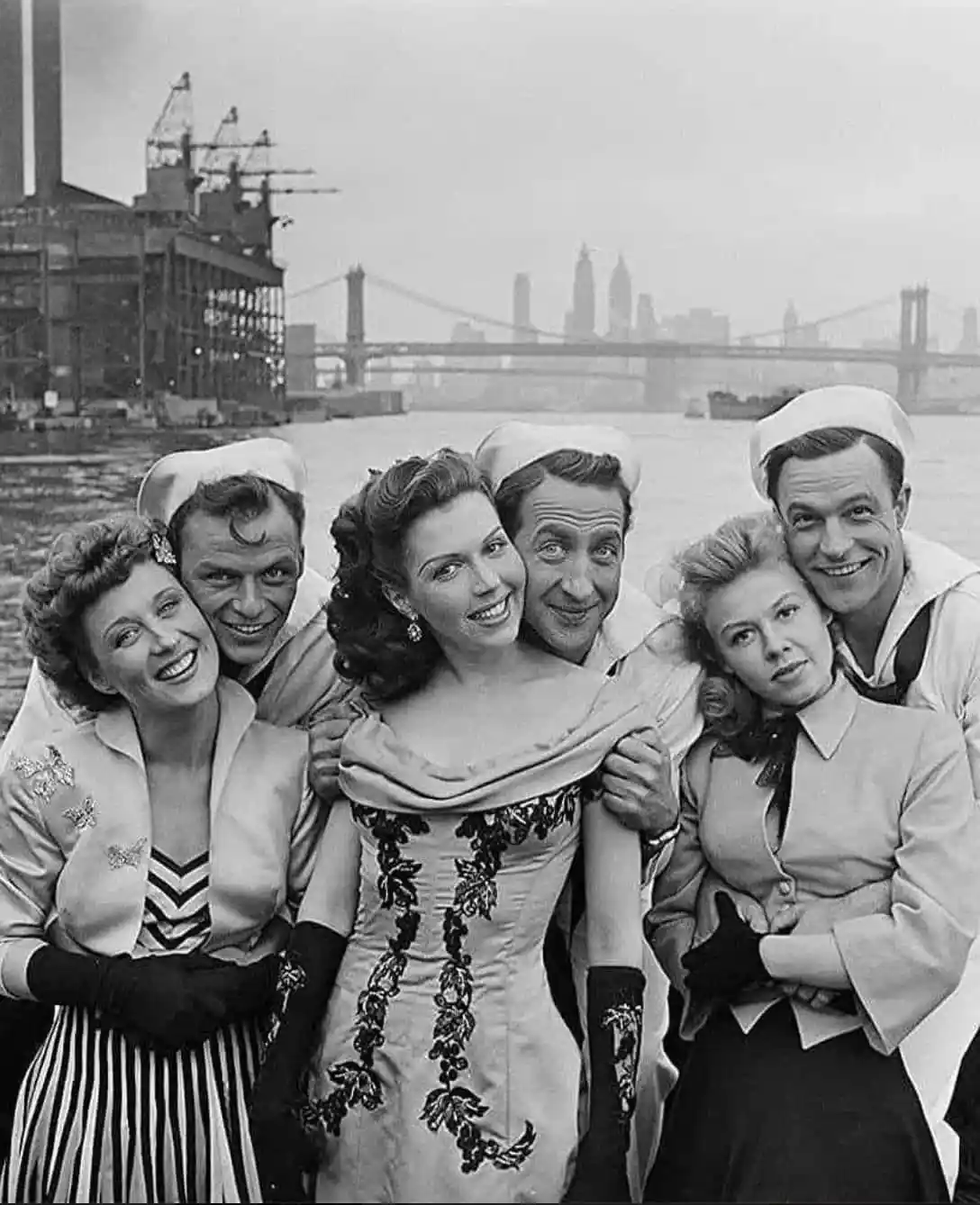 A black-and-white photo of five actors posing together on a boat with a city skyline and bridge in the background. Four men…