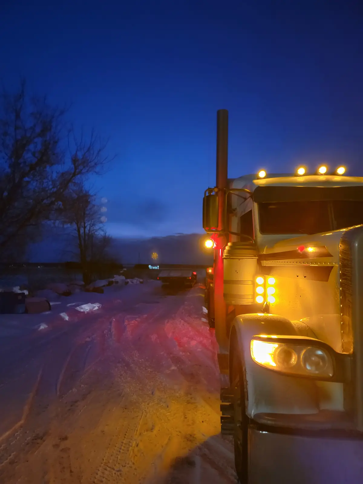 A large white semi-truck is parked on a snowy road at dusk, its bright yellow headlights and red clearance lights…