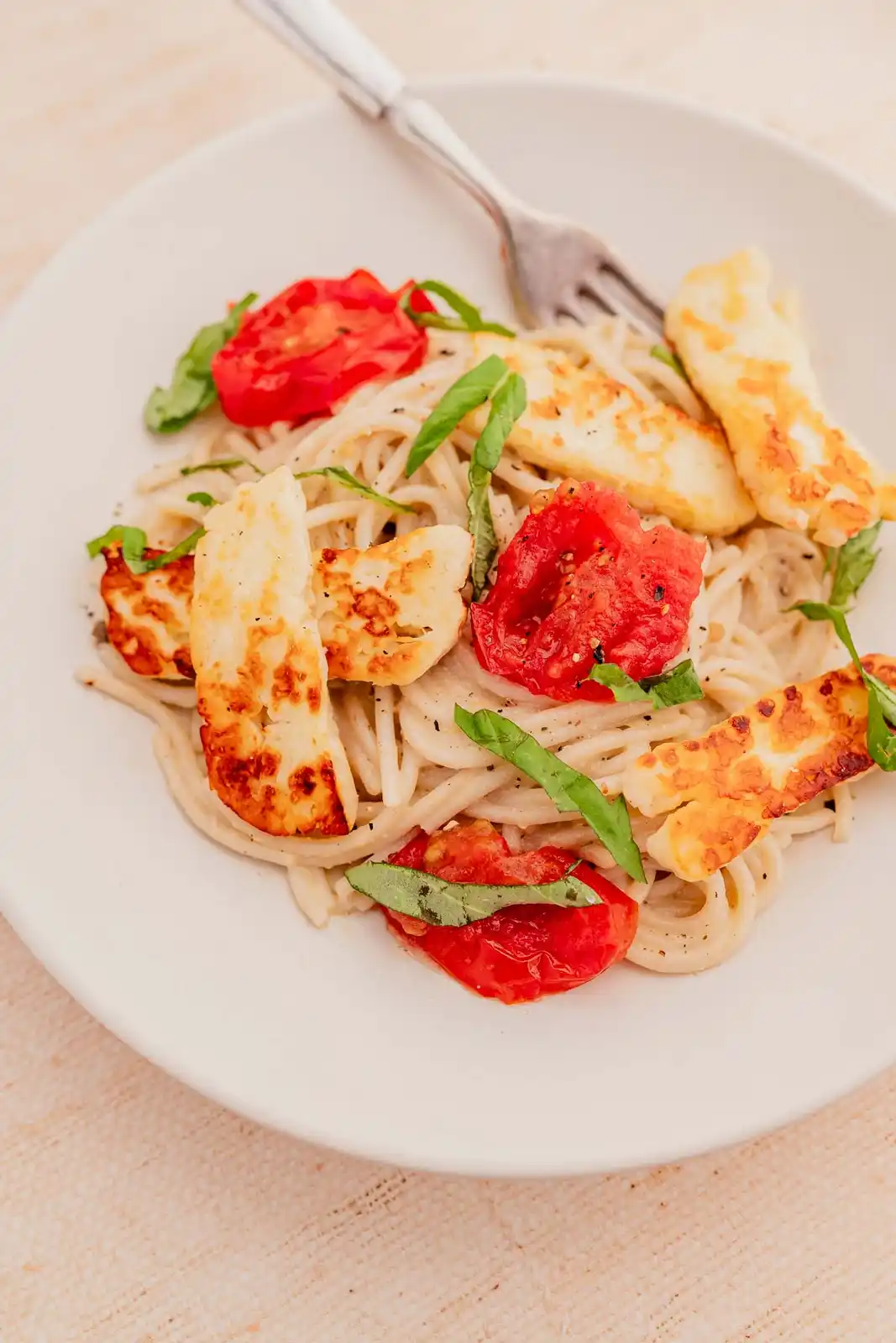 A close-up photo shows a white plate of pasta topped with golden-brown halloumi cheese slices, roasted red tomatoes, and…
