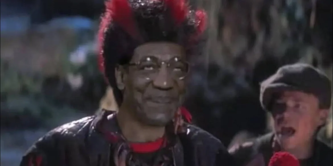 A man with a red and black mohawk wig and glasses smiles while wearing a black leather jacket over a red shirt. Behind him…