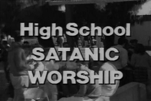 A grainy black-and-white image shows a group of people seated at tables in what appears to be a school cafeteria or similar…