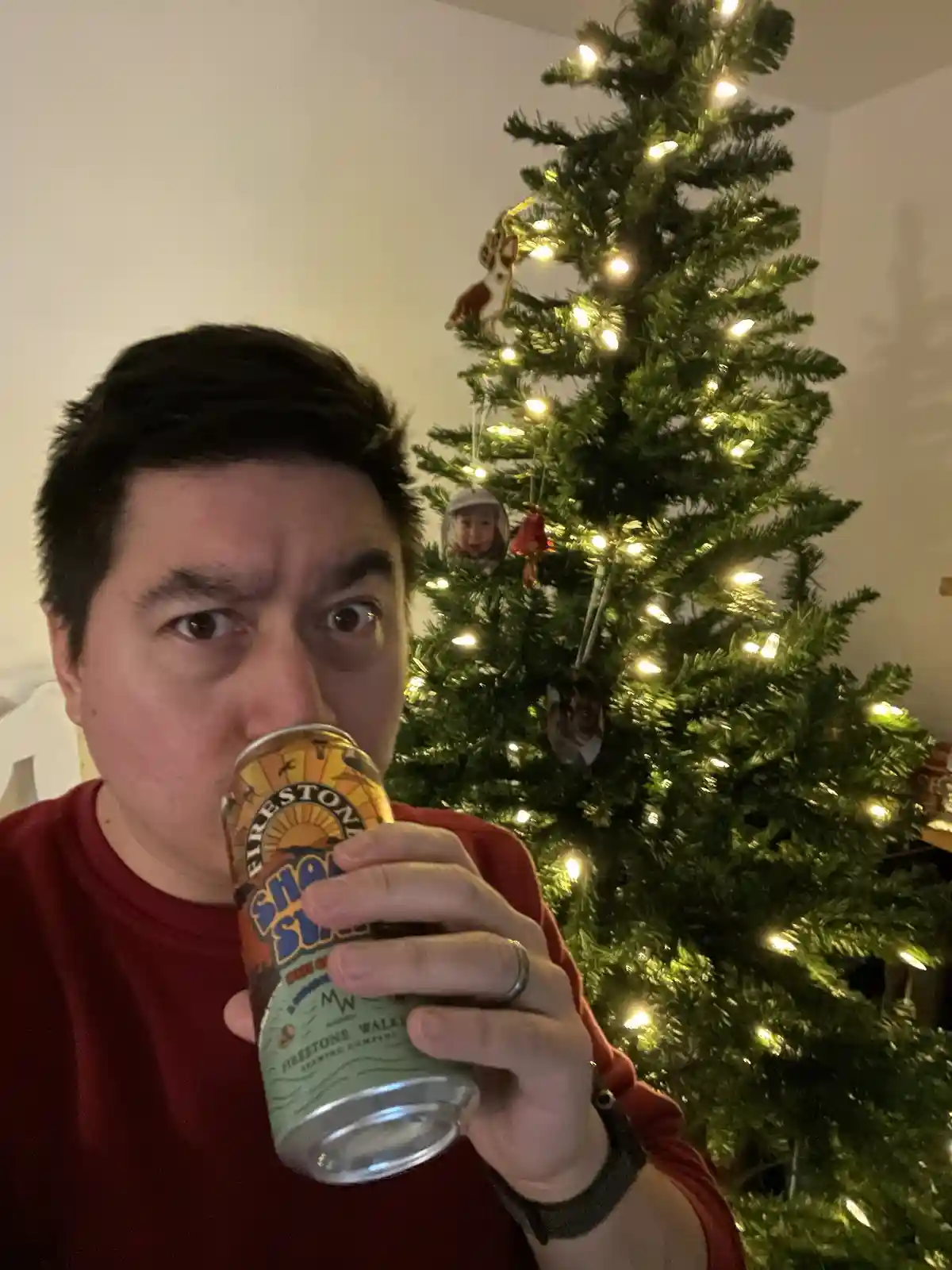 A man in a red shirt holds a can of Firestone Walker 'Sunset' beer up to his mouth, looking directly at the camera with…