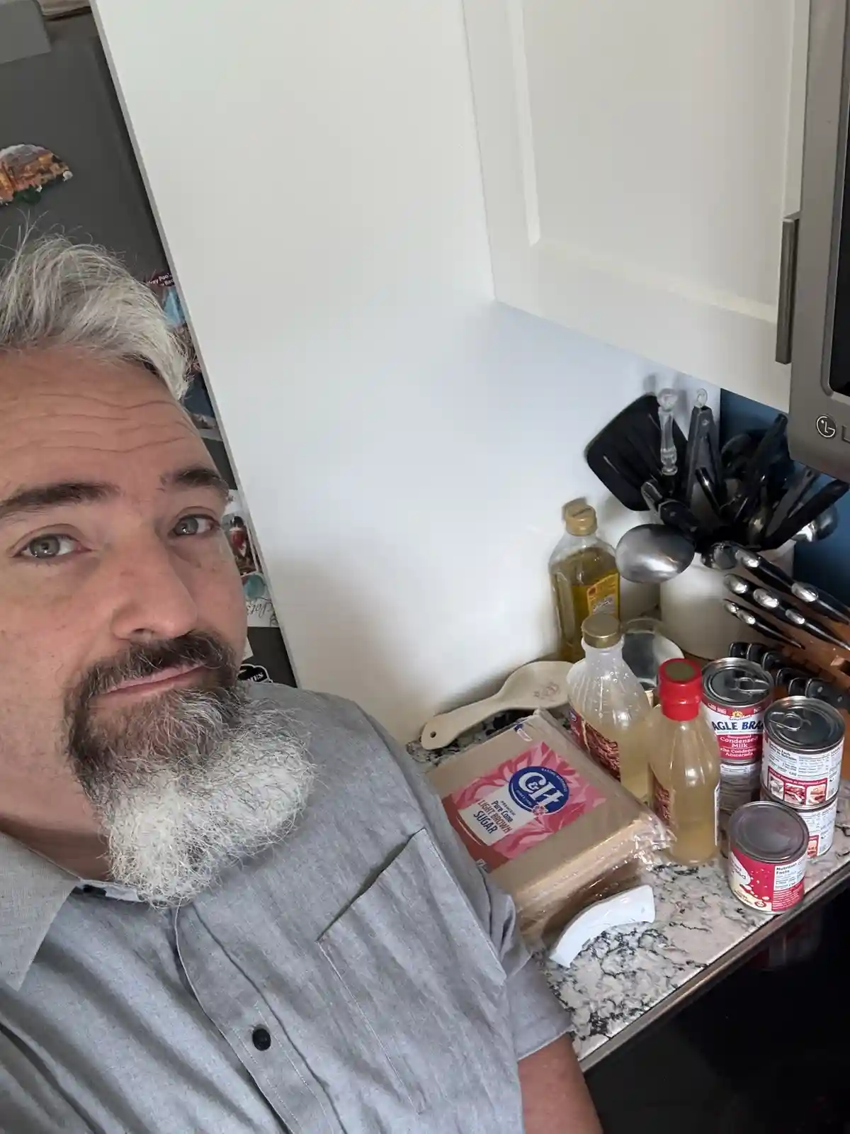 A man with a gray beard and mustache looks directly at the camera with a smug expression while standing in a kitchen. Behind…