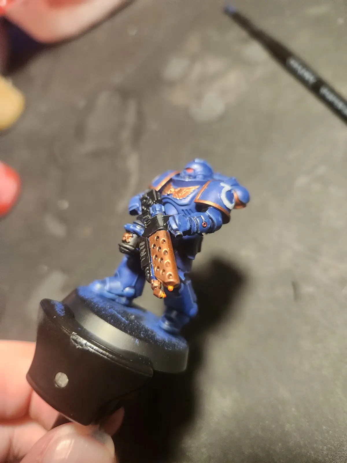 A hand holds a painted blue and gold miniature of a Space Marine from Warhammer 40,000, showing off its detailed armor and…