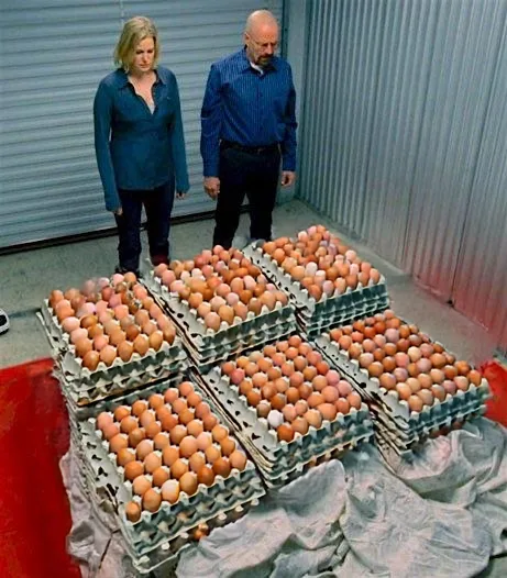 A man and a woman stand in a storage unit looking down at a large pile of egg cartons filled with brown eggs. The woman has…