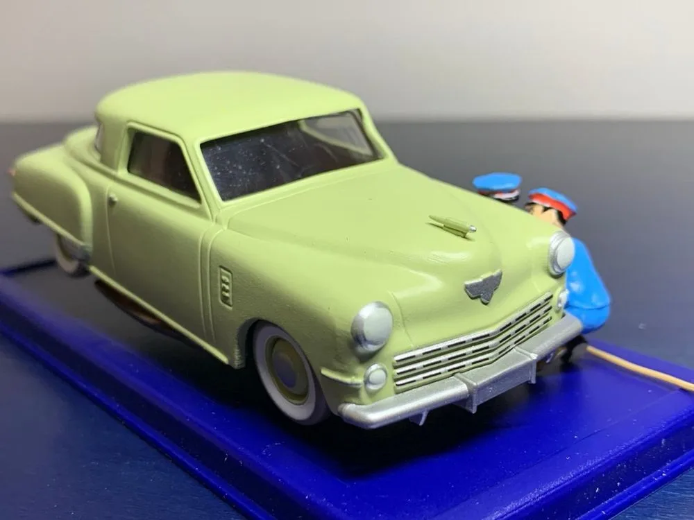A pale green vintage toy car sits on a blue display base, with a small toy police officer figurine leaning against its front…