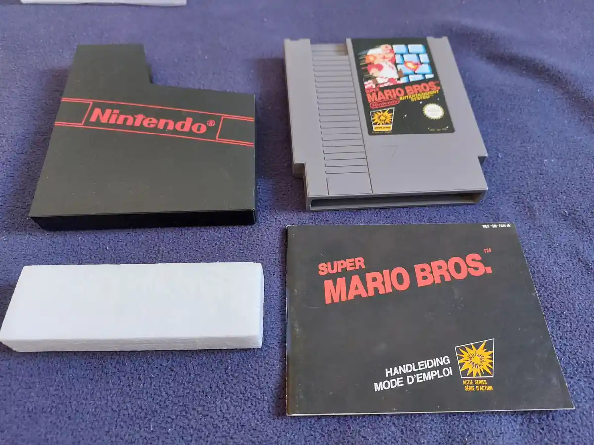 A photo of a Super Mario Bros. NES game cartridge, its black box, and instruction manual laid out on a blue carpet. The…