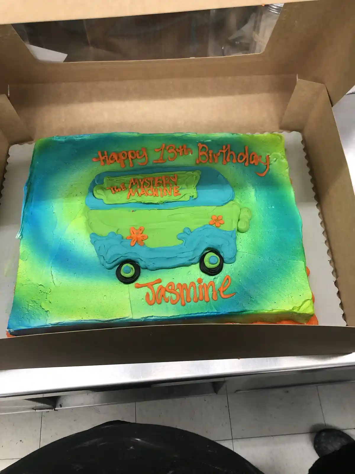A rectangular birthday cake in a cardboard box features a frosting design of the green and blue Mystery Machine van from…