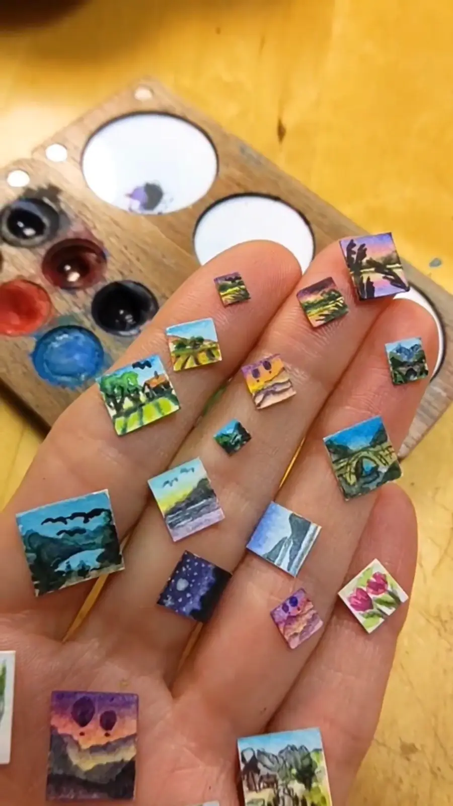 A hand with fingernails painted with miniature watercolor landscapes, including mountains, sunsets, flowers, and night…