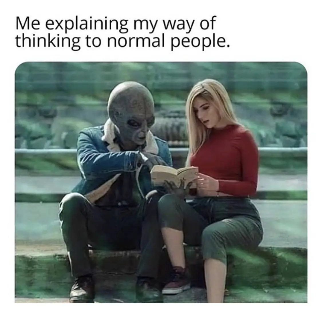 An alien with a large head and dark skin sits beside a human woman with blonde hair, both looking at an open book. The alien…