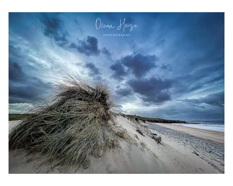 A dramatic landscape photograph of a windswept beach with a large dune covered in grass in the foreground, under a moody…