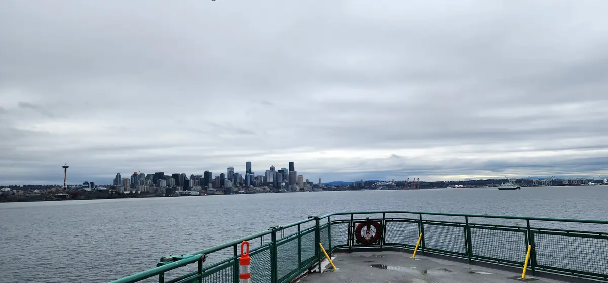 A photo taken from a ferry deck shows the Seattle skyline under an overcast sky, with the Space Needle visible on the left…