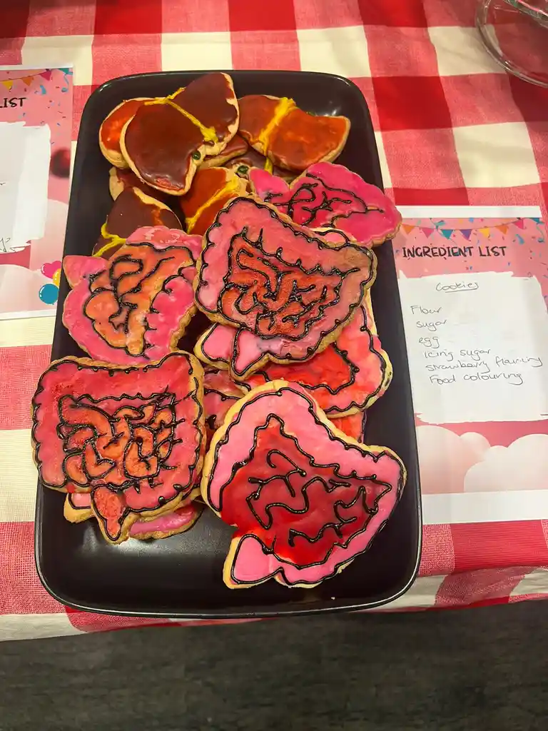 A black tray holds heart-shaped cookies decorated with pink icing and dark chocolate outlines resembling brain or organ…