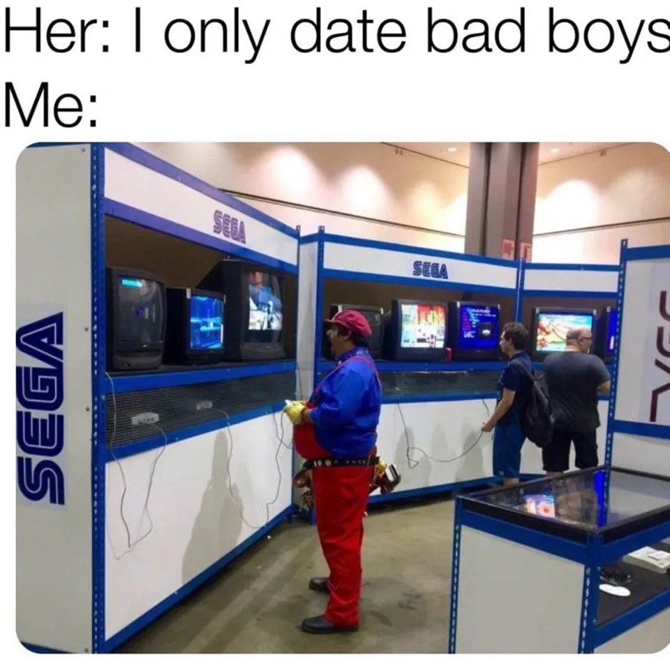 A man dressed in a Mario costume stands at a Sega arcade game booth, holding a controller, while other people play games…