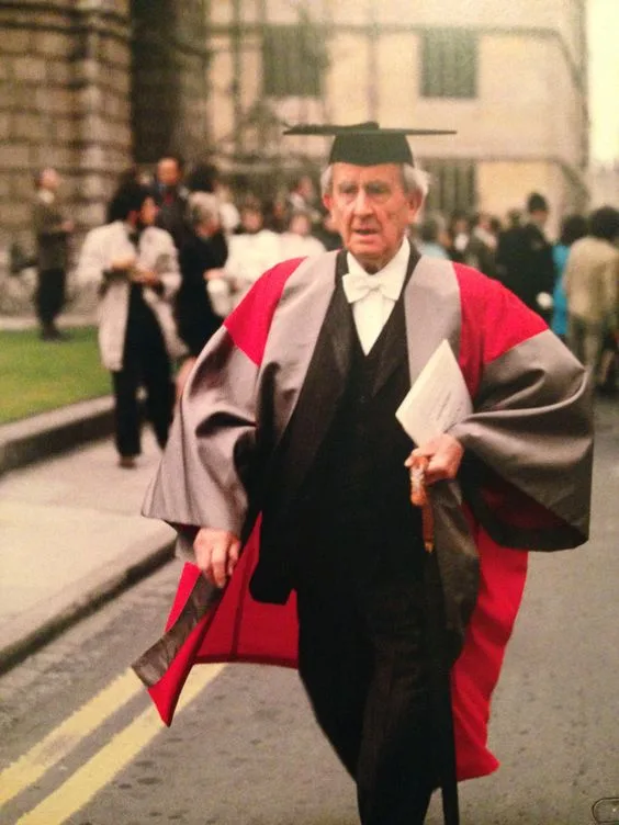 An older man in academic regalia, including a black graduation cap and a red-and-grey robe, walks outdoors while holding a…