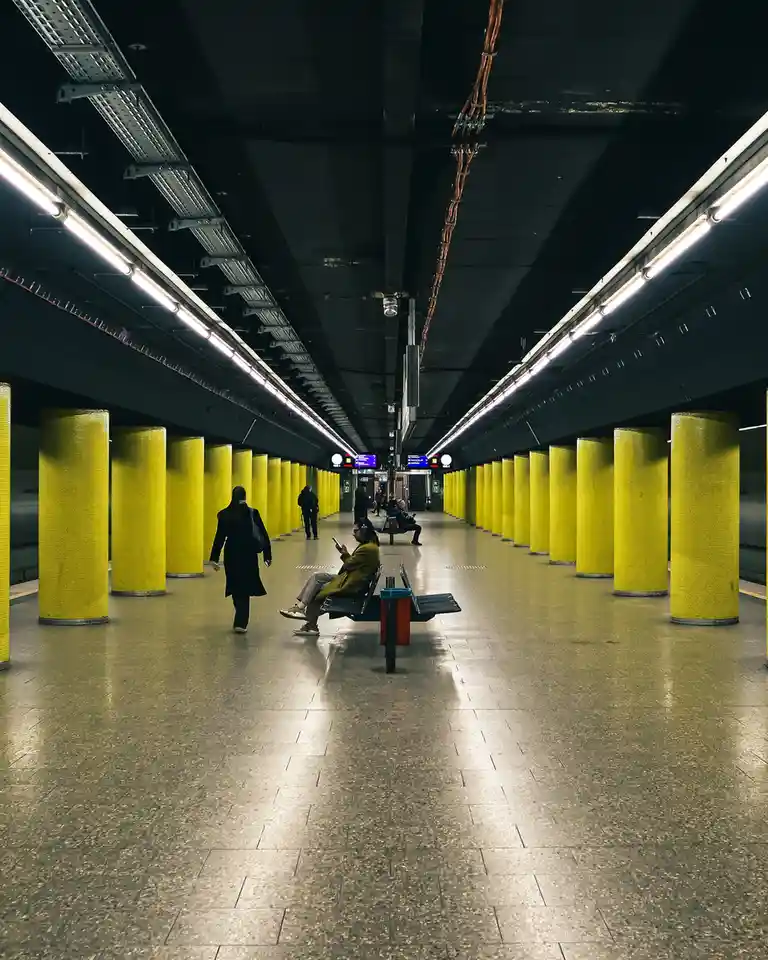 A wide-angle shot of a subway station with rows of bright yellow pillars, fluorescent lighting, and a few people. A person…