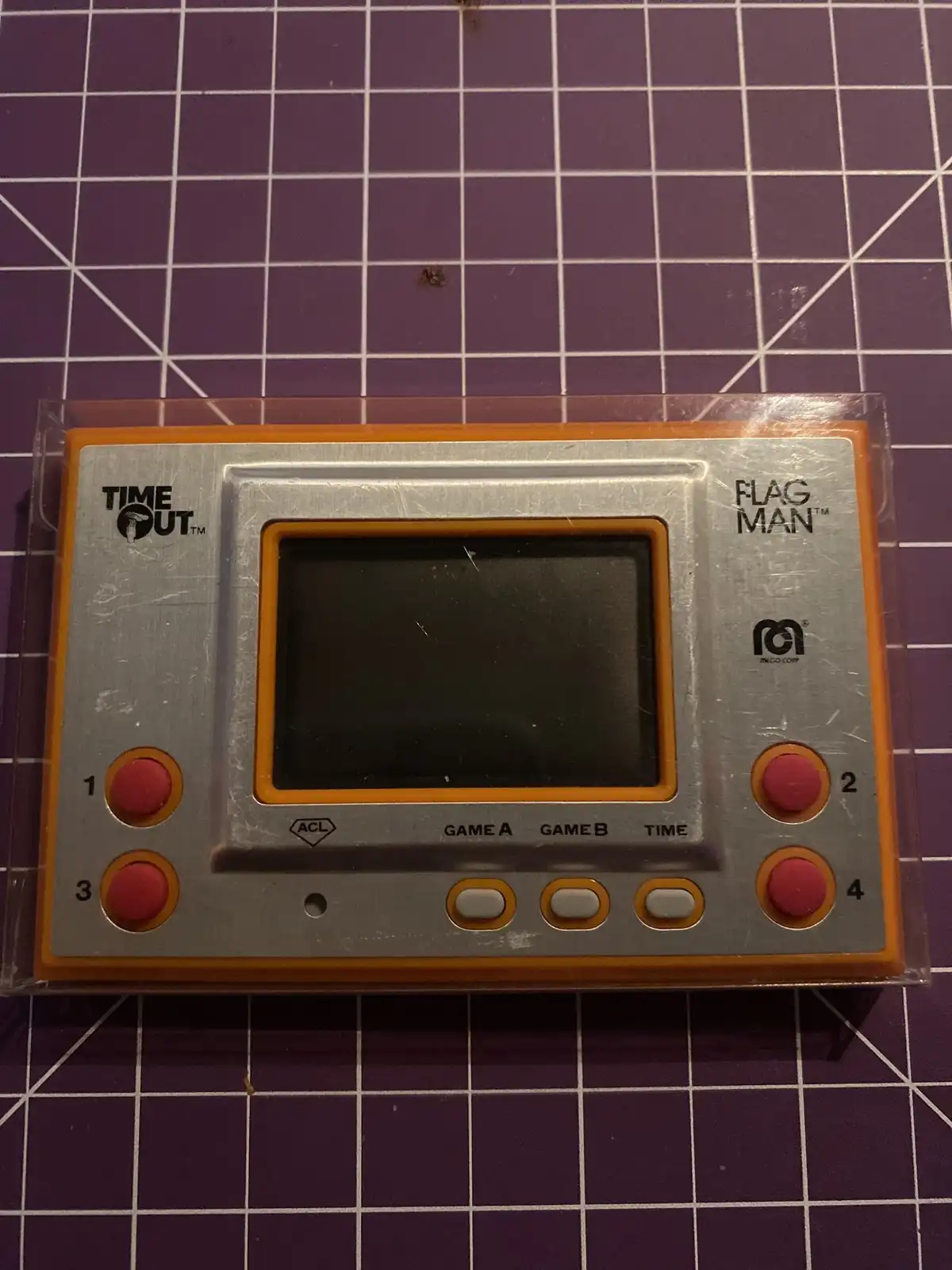 A silver and orange handheld game console labeled 'TIME OUT™' and 'FLAG MAN™' by Mego Corp, featuring four red buttons…