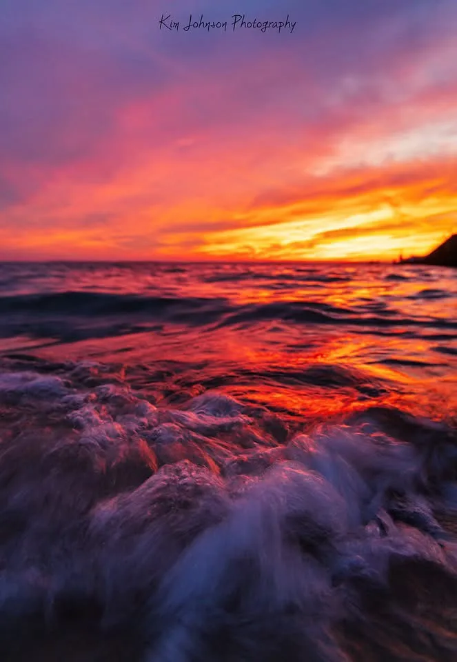 A vibrant, blurred photograph of ocean waves crashing at sunset, with fiery orange and purple skies reflected in the water…