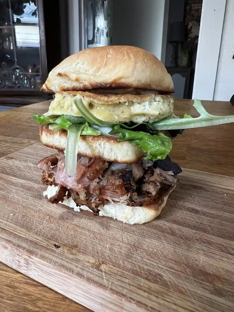 A towering, messy sandwich made with a toasted bun, crispy bacon, shredded meat, lettuce, and a fried egg, sits on a wooden…