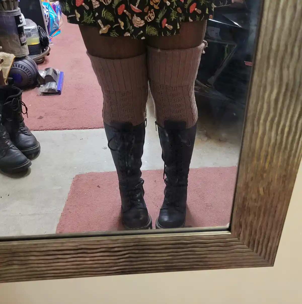 A mirror selfie shows a person wearing brown ribbed knee-high socks and black lace-up knee-high boots, standing on a pink…