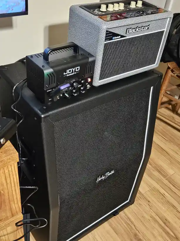 A Blackstar guitar amp sits on top of a Harley Benton speaker cabinet, with a small black JOYO banampP.xl device placed on…