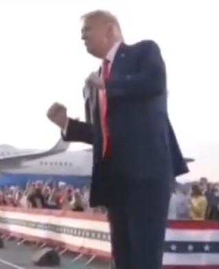a man in a dark suit and red tie stands on a red, white, and blue striped platform with his fist raised, facing away from…