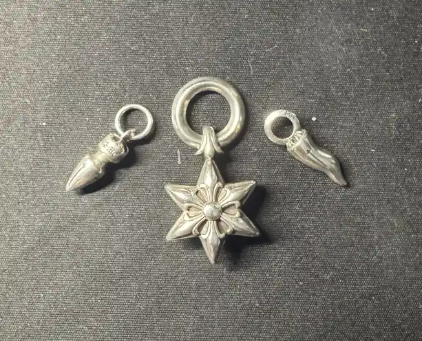 Three silver charms lie on a dark gray fabric: a central star-shaped pendant, a pointed bullet-shaped charm on the left, and…