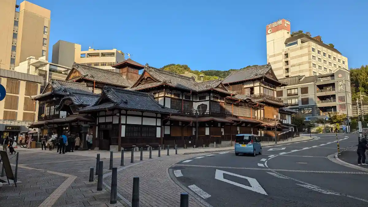 A wide-angle photo of a traditional Japanese wooden building with dark tiled roofs and white walls, situated on a paved…
