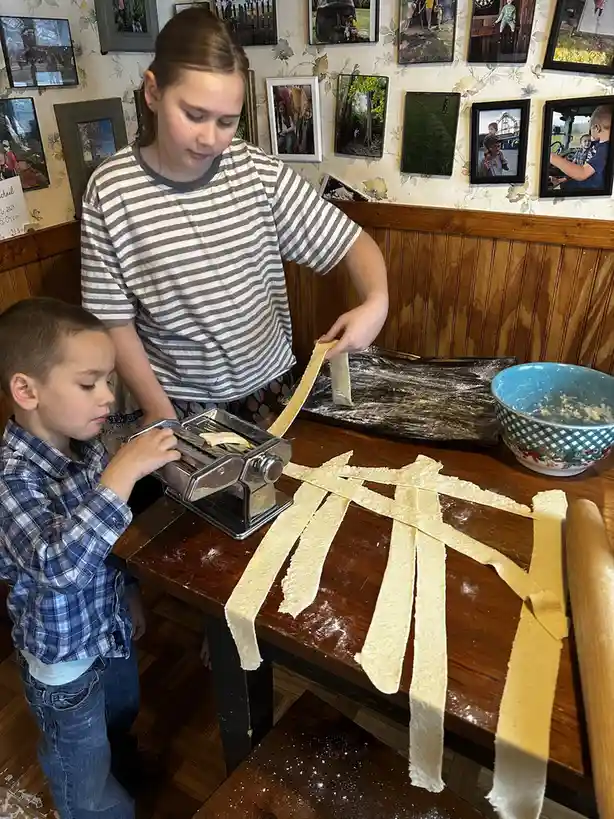 Two children, a girl in a striped shirt and a boy in a plaid shirt, are making pasta using a manual pasta maker on a wooden…