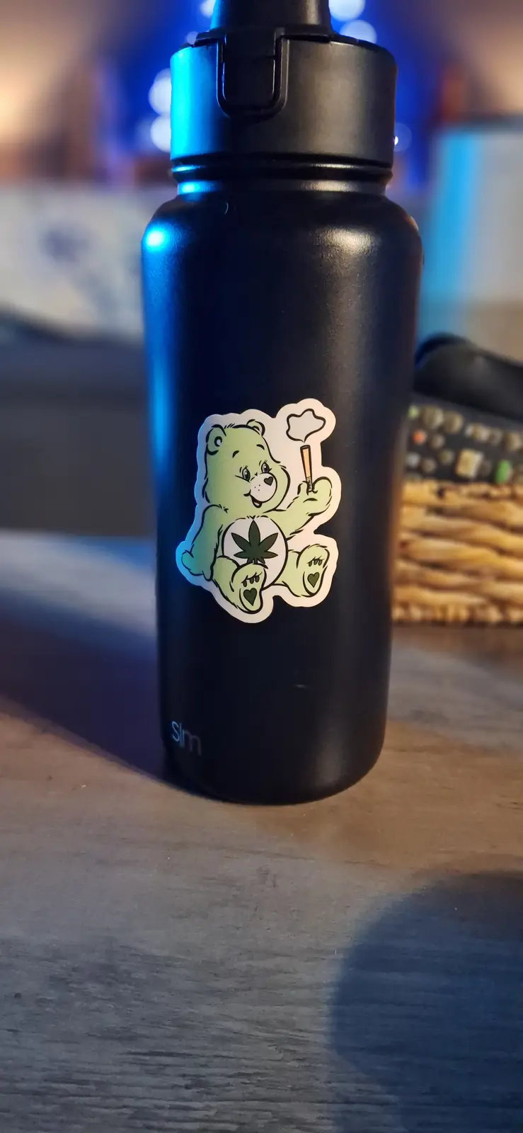 A black water bottle with a sticker of a green Care Bear holding a cannabis leaf and smoking a joint, set against a blurred…