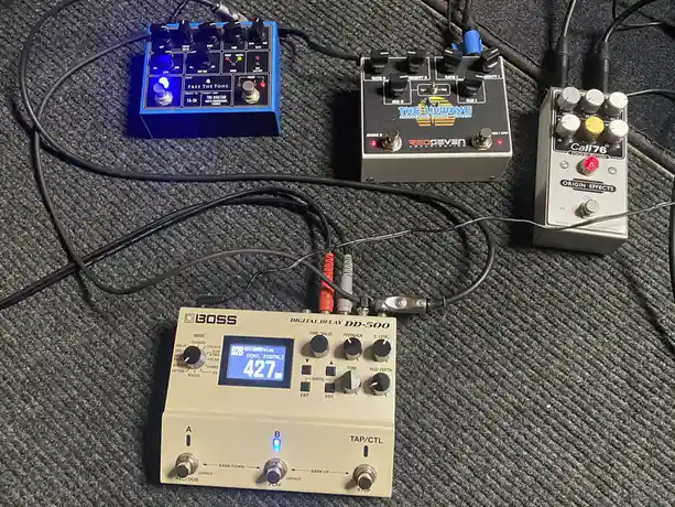 A photo of four guitar effects pedals connected by cables on a grey carpet, with the Boss DD-500 digital delay pedal in the…