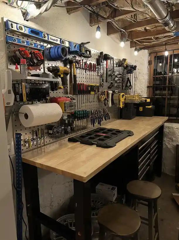 A well-organized basement workshop features a wooden workbench with black drawers, a pegboard wall densely packed with…