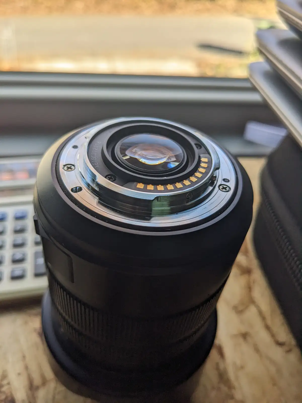 A close-up photo of a black camera lens sitting on a wooden surface, with a calculator and window visible in the background…