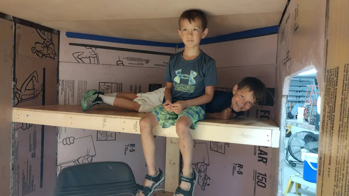 Two young boys are inside a makeshift bunk bed structure made of wooden planks and pink insulation boards with instructional…