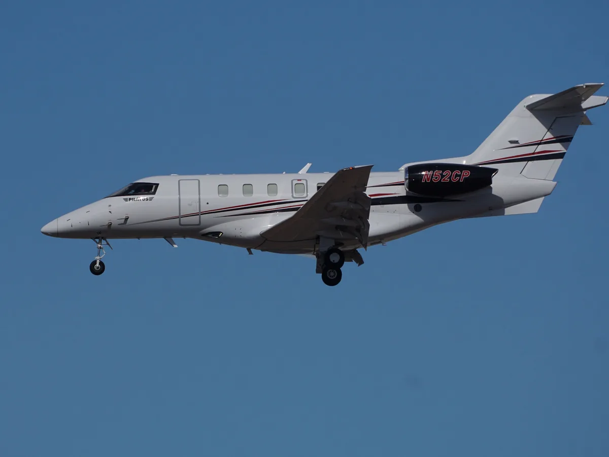 A white Pilatus business jet with red and black stripes is flying against a clear blue sky, its landing gear down. The…