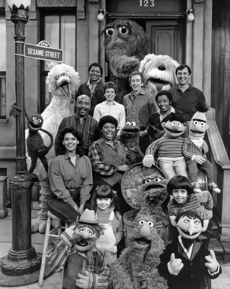 A black-and-white group photo of the original Sesame Street cast and puppets posing in front of 123 Sesame Street, including…