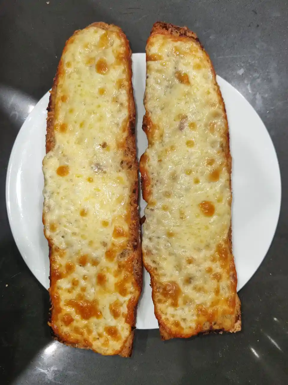 Two long, toasted bread slices topped with melted cheese sit side by side on a white plate, placed on a dark countertop. The…