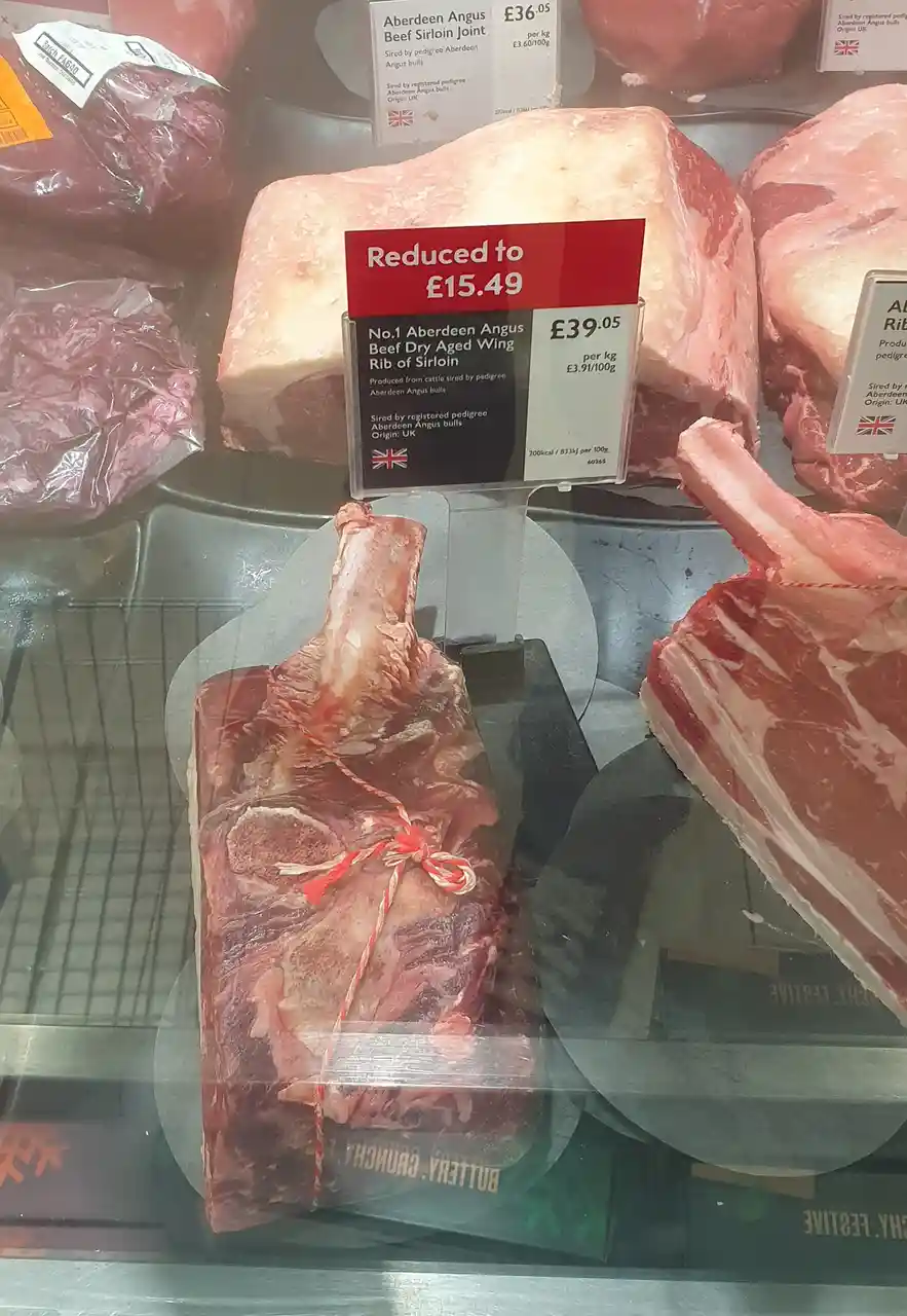 A close-up of a beef rib of sirloin in a supermarket meat display, with a red price tag stating 'Reduced to £15.49' over the…