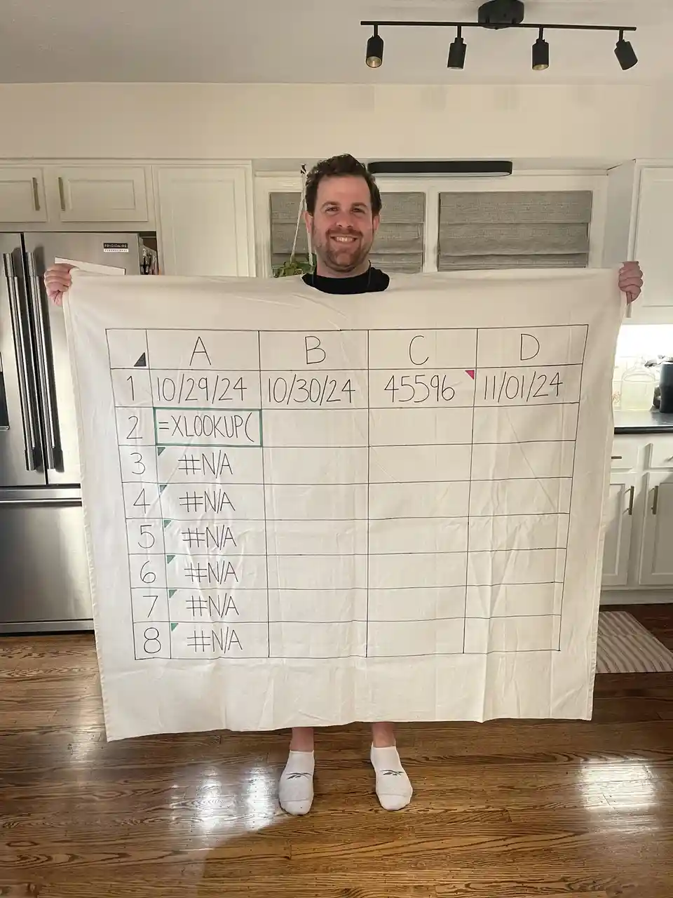 A man in a kitchen holds up a large white blanket with a hand-drawn spreadsheet grid. The grid has columns labeled A, B, C…