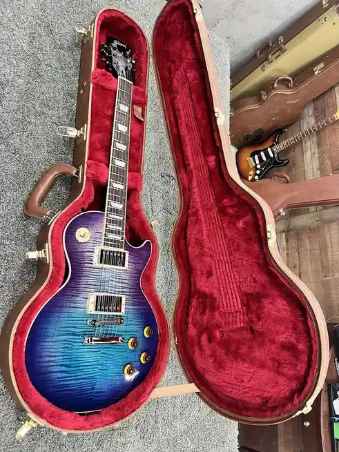 A Gibson Les Paul electric guitar with a vibrant blue-to-purple gradient finish sits inside an open, empty brown hardshell…