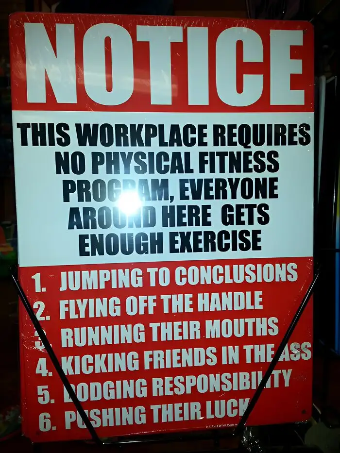 A red and white workplace notice sign humorously claims that no physical fitness program is required because everyone gets…