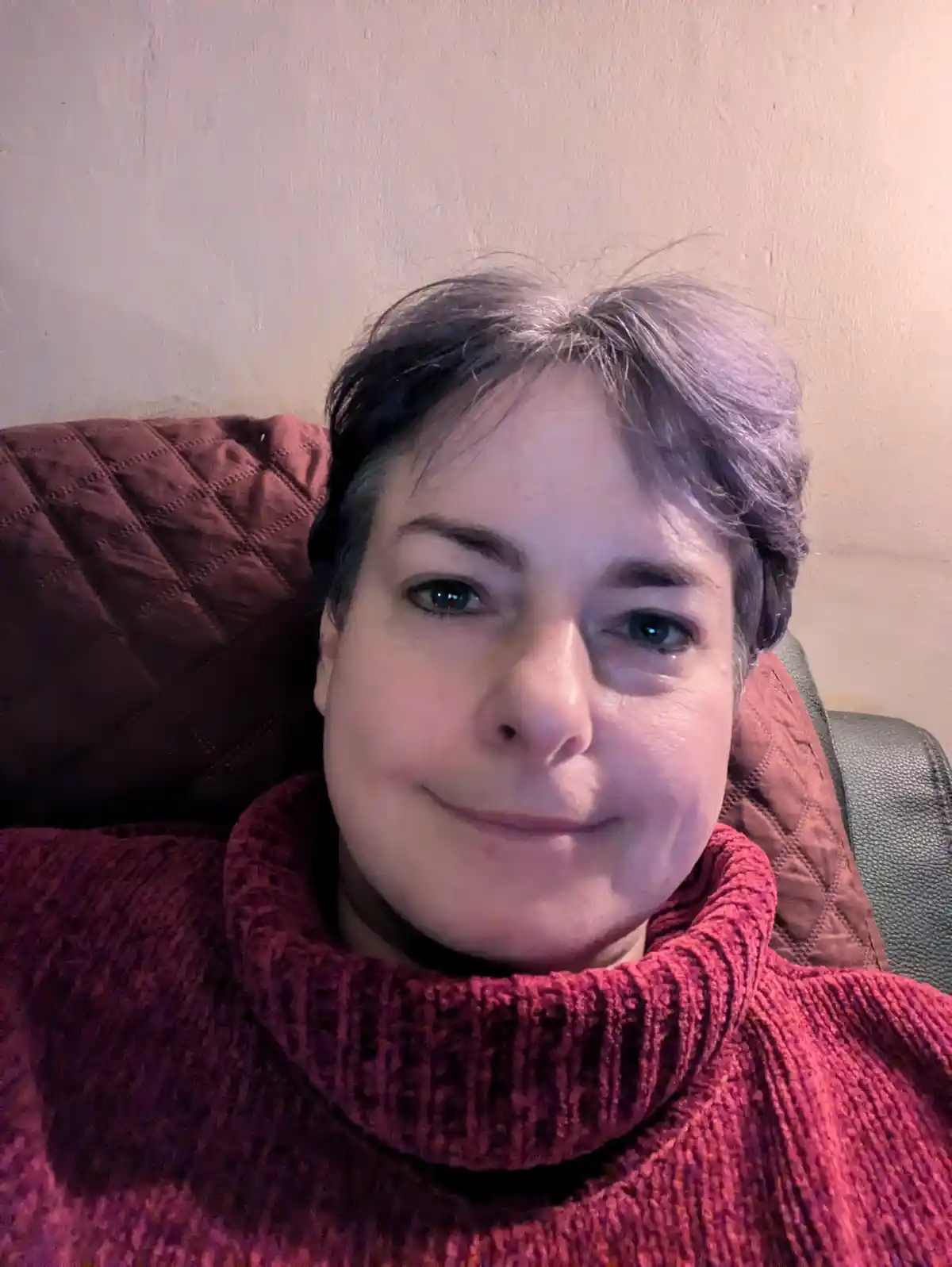 A woman with short grayish hair and green eyes smiles gently while wearing a red turtleneck sweater, seated against a brown…