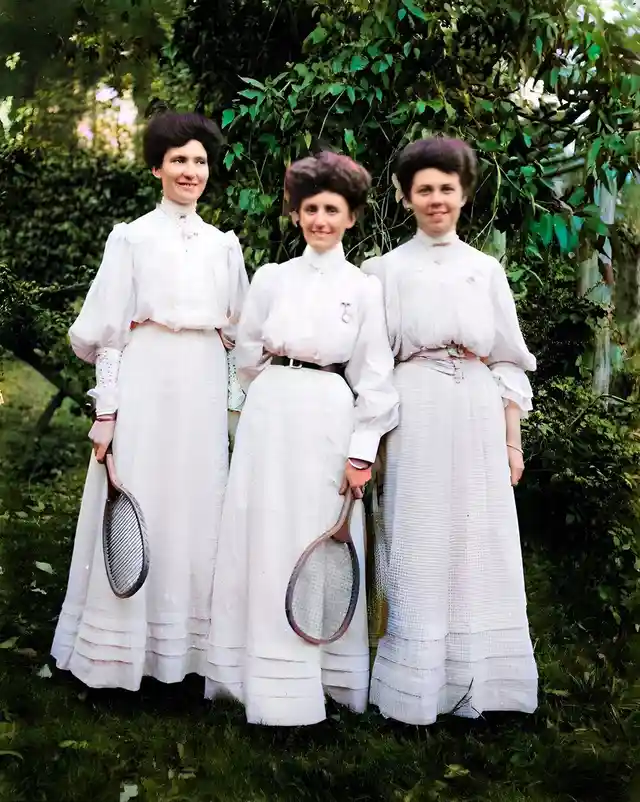 Three women in vintage white tennis dresses from the early 1900s stand outdoors holding old-fashioned wooden tennis rackets…